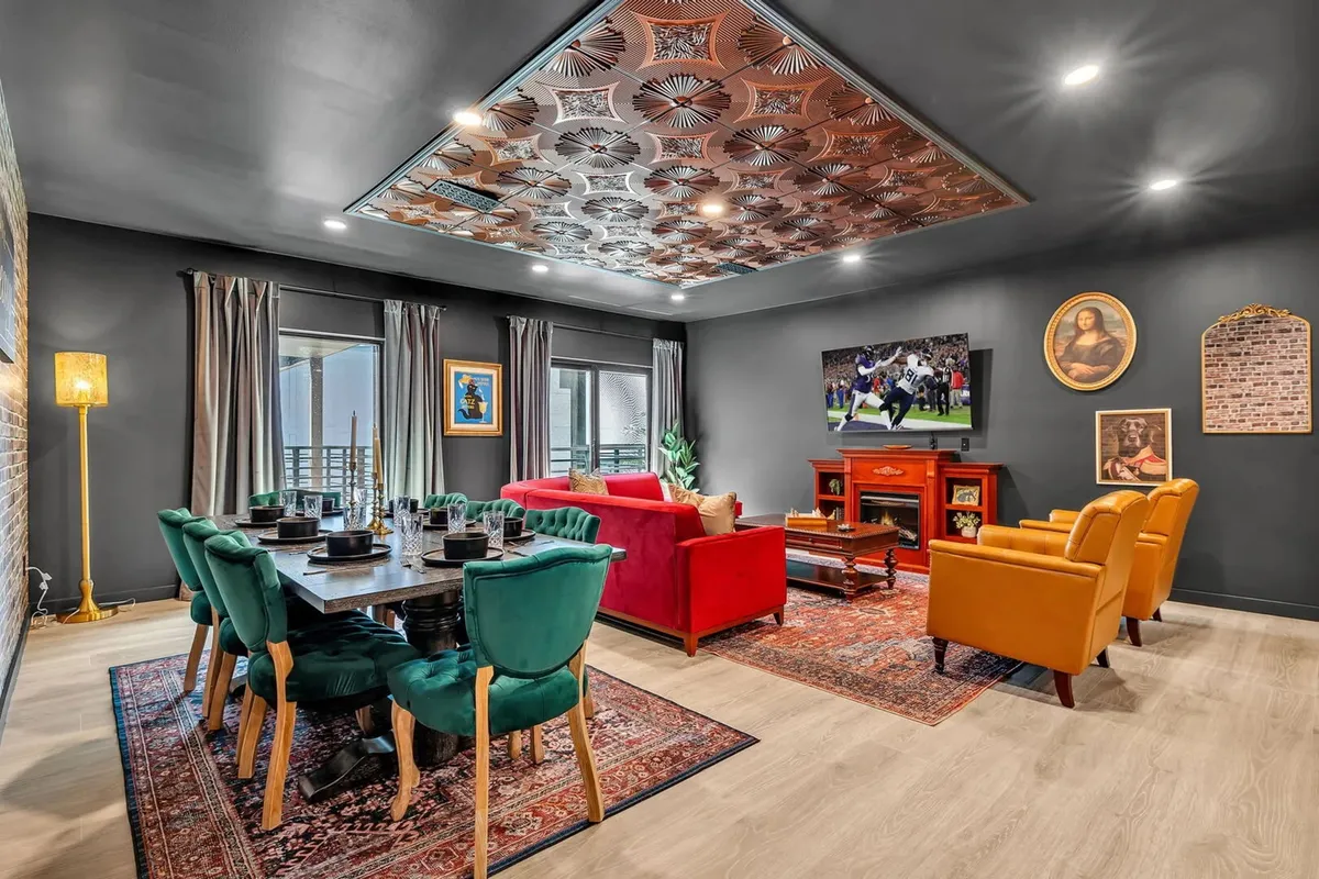 The hero image showcases a vibrant, open-plan living and dining area bathed in natural light. A sleek dining table is set for gatherings, surrounded by a bold red sofa and orange armchairs, all accented by a decorative ceiling. The TV displays a sports broadcast, setting a lively, communal atmosphere ideal for group celebrations or relaxing evenings.