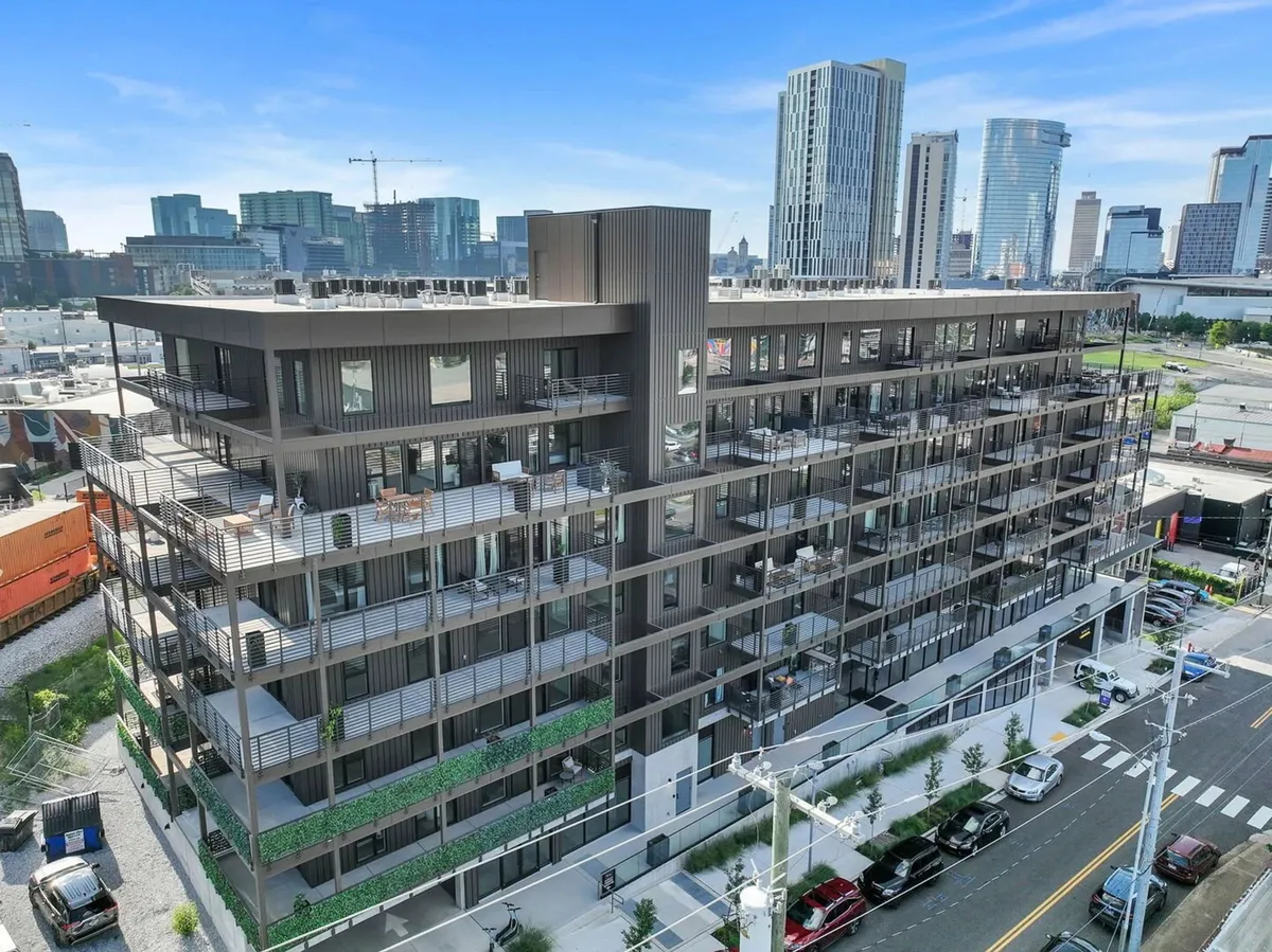 A wide exterior shot of the modern apartment building, with a rooftop terrace and balconies adorned with greenery, set against the urban Nashville skyline. The rooftop area invites guests to socialize while taking in panoramic city views.