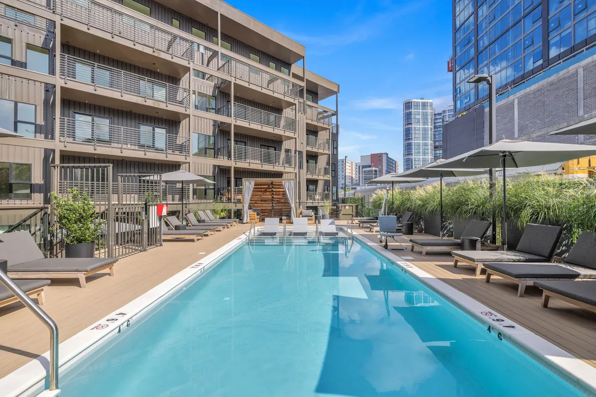 A sparkling outdoor swimming pool is the central focus, surrounded by comfortable lounge chairs and lush greenery, with the building’s modern facade in the background. The setting suggests a vibrant place for sunbathing, swimming, or relaxing poolside during warm months.
