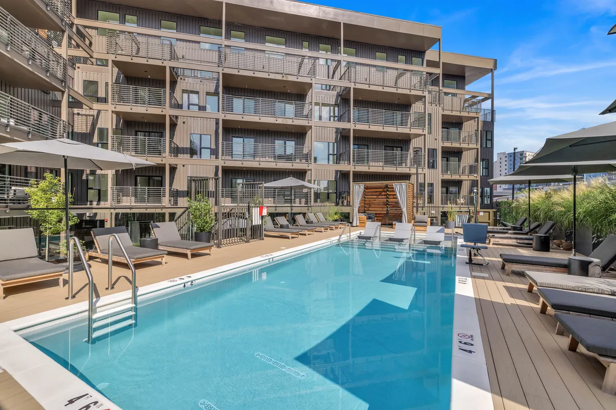 A bright exterior scene featuring a sparkling swimming pool surrounded by lounge chairs and umbrellas, set against the contemporary facade of the apartment building. The area is designed for relaxation and socializing, with ample space for guests to unwind or gather for poolside parties.