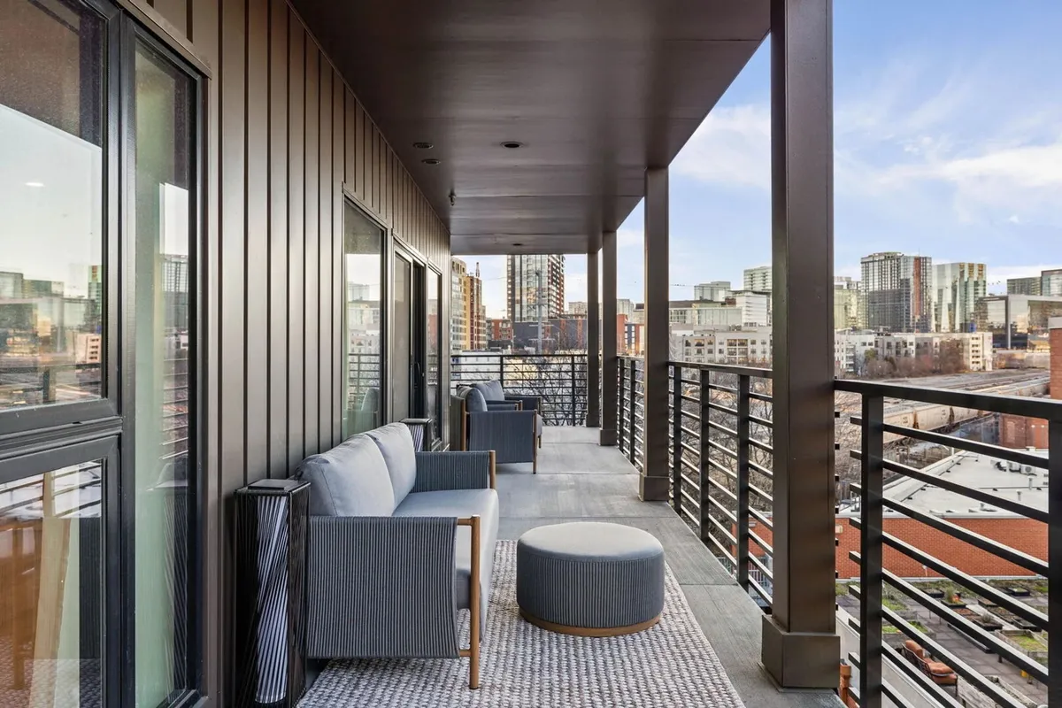 A serene outdoor seating area on a balcony or terrace with views overlooking the city skyline. The setup includes comfortable seating and a textured rug, inviting guests to unwind while enjoying the urban panorama.