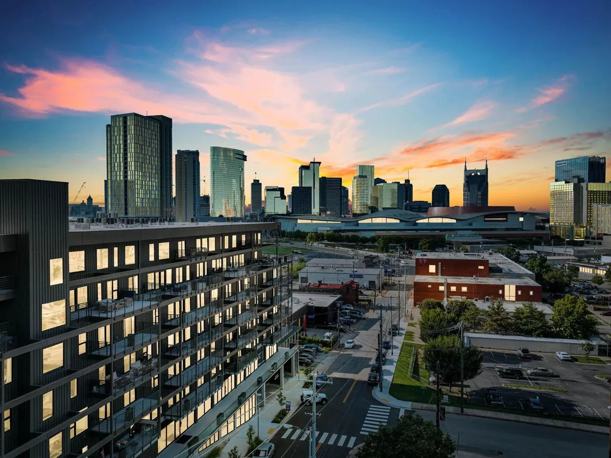 A wide view of a modern apartment building set against the Nashville city skyline, with the warm glow of sunset coloring the sky. The urban setting emphasizes proximity to downtown, capturing the feeling of city exploration and adventure.