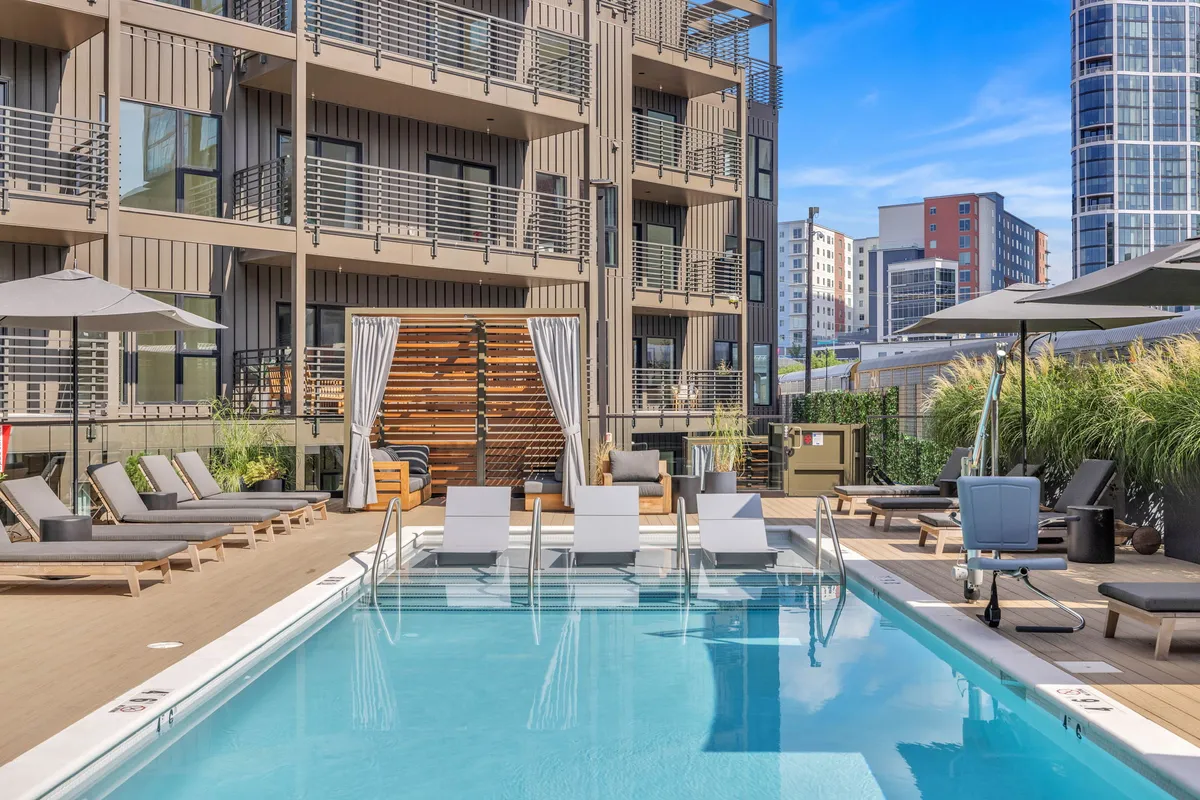 A vibrant outdoor swimming pool area with lounge chairs, umbrellas, and the building facade in the background. The space is designed for relaxation and socializing, with plenty of seating and shade for families to enjoy.