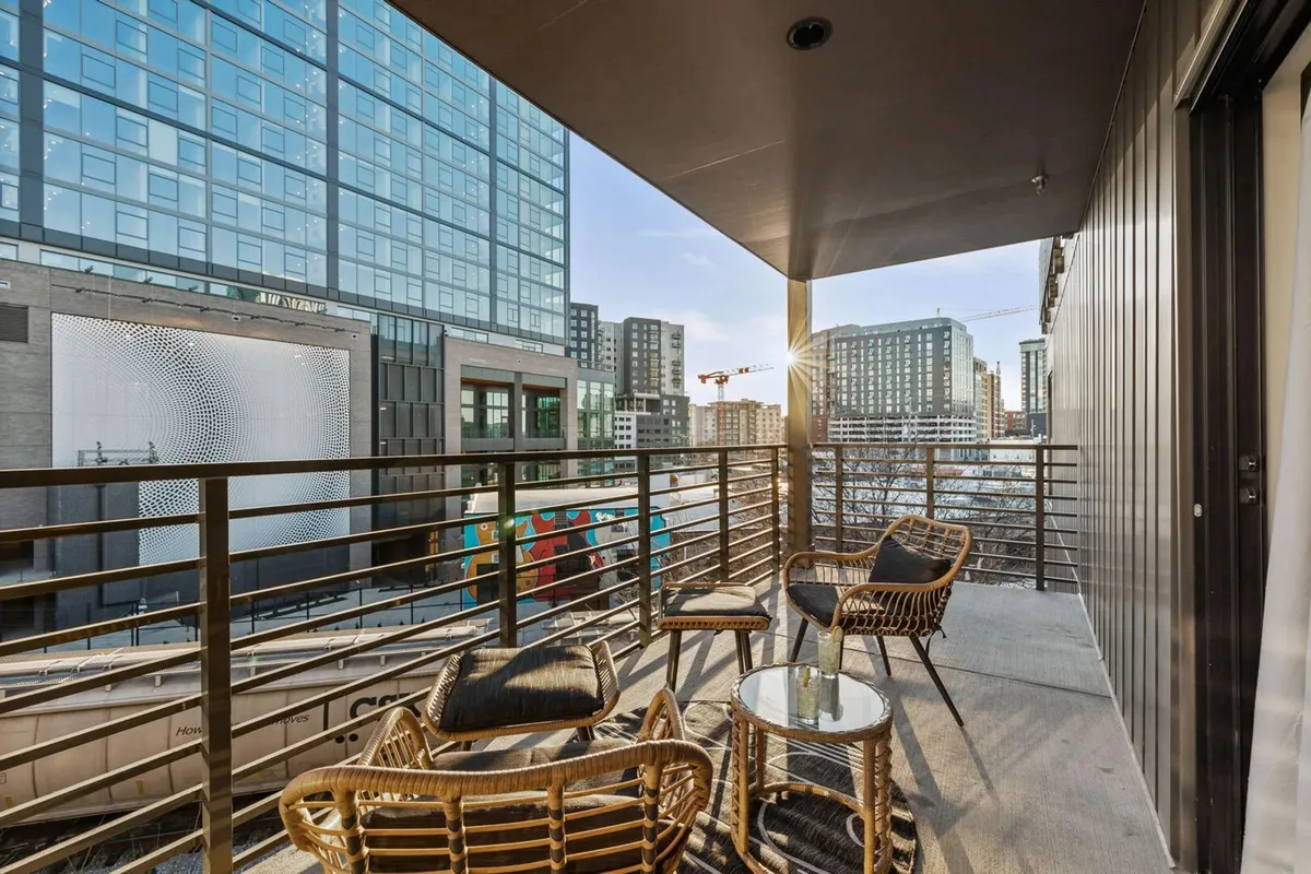 A balcony furnished with comfortable chairs, offering a vibrant view of the city skyline and an art mural on a neighboring building. The atmosphere is contemporary, capturing the energy of urban life from a private vantage point.