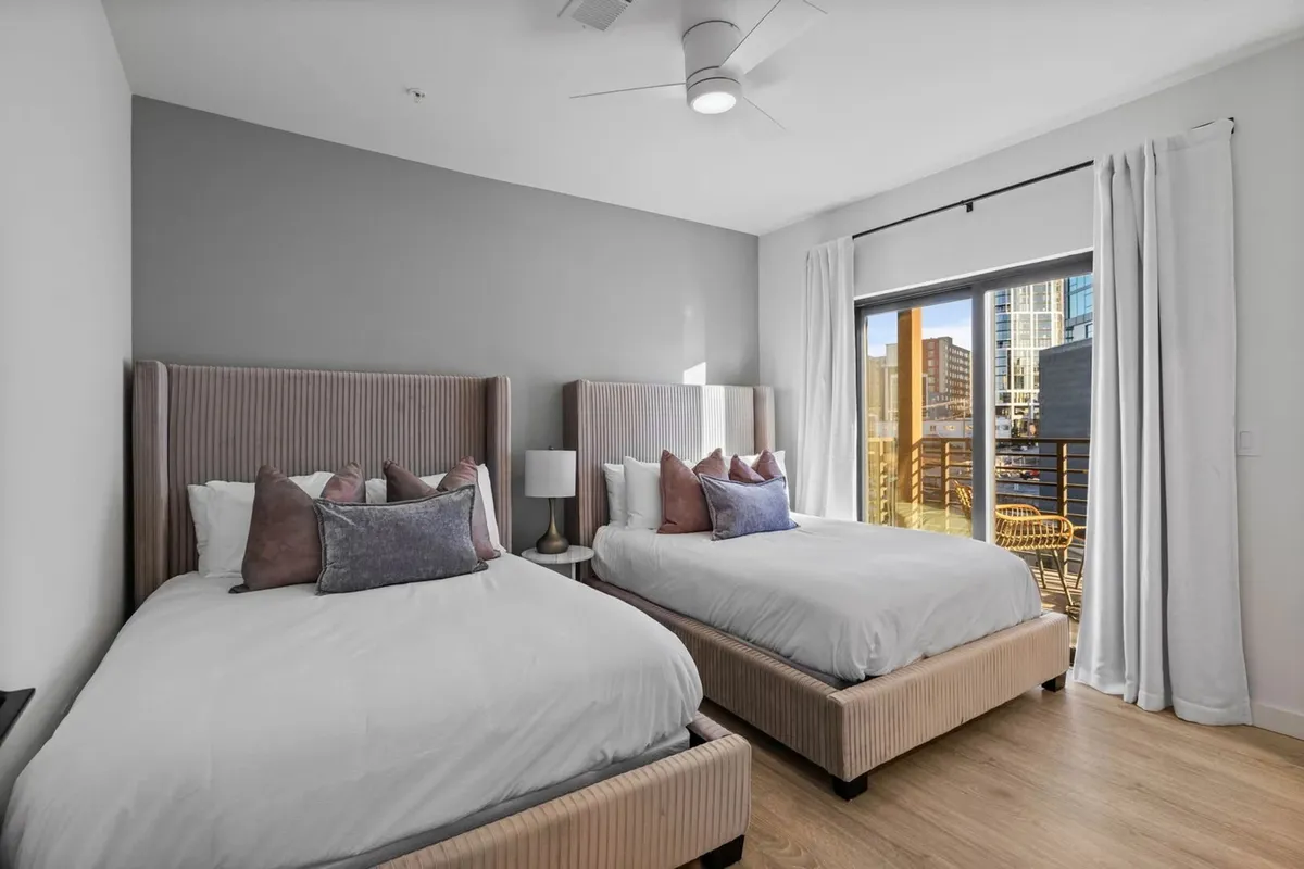 A cozy bedroom with two beds featuring upholstered headboards, a large window streaming in natural light, and decorative pillows accentuating the inviting mood. The soft palette and thoughtful arrangement set a tranquil tone ideal for relaxation and togetherness.