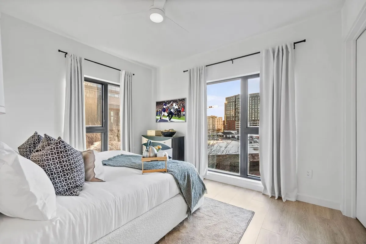 This image presents a modern bedroom with a plush bed adorned with decorative pillows, a large window offering a city view, and a wall-mounted television displaying a sports event. The nightstand features tasteful decor, and the overall atmosphere is both inviting and contemporary.