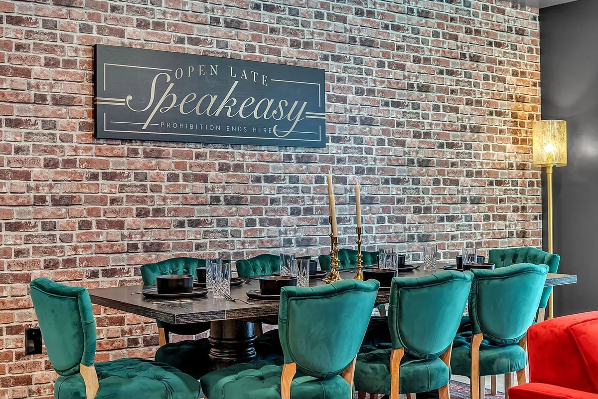 A stylish dining area with a rustic brick wall, an elegant dining table surrounded by plush teal velvet chairs, a glowing speakeasy sign, and flickering candles sets the mood for celebratory dinners or intimate gatherings. The ambiance is sophisticated yet comfortable, ideal for memorable moments with friends and family.